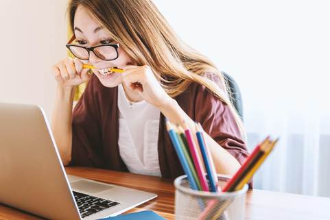 A young woman sitting at a desk with a laptop, looking anxious while biting on a pencil, surrounded by colorful pens in a cup.