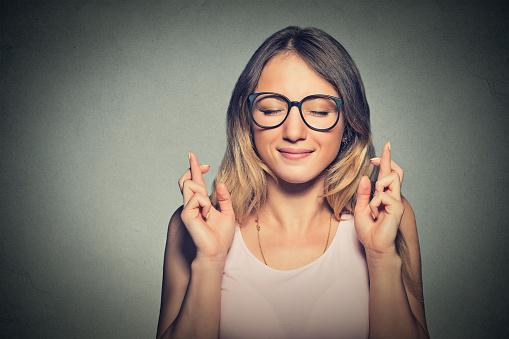 A young woman with glasses smiling and crossing her fingers, symbolizing hope and good luck.