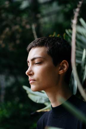 A person with short hair peacefully meditating among green plants, eyes closed, conveying a sense of calm and mindfulness.