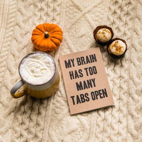 A cozy scene featuring a cup of coffee topped with whipped cream, a small decorative pumpkin, and chocolate cupcakes placed on a knitted blanket, alongside a note that reads 'MY BRAIN HAS TOO MANY TABS OPEN.'