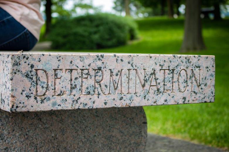 A close-up of a stone bench with the word 'DETERMINATION' engraved on it, set in a green park environment.
