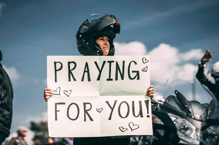 A person wearing a motorcycle helmet holds a sign that reads 'PRAYING FOR YOU!' while standing outdoors.