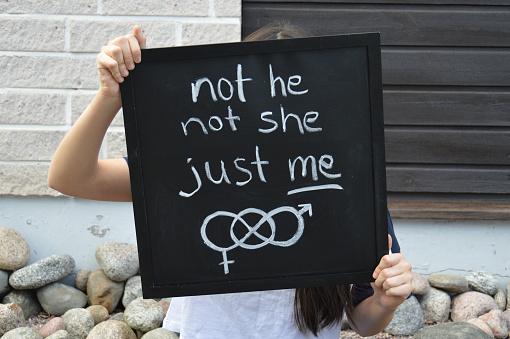 A person holding a chalkboard sign that reads 'not he not she just me', symbolizing gender fluidity and self-identity, with gender symbols in the background.