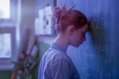 A young woman with her head resting against a chalkboard covered in math equations, appearing overwhelmed or frustrated.