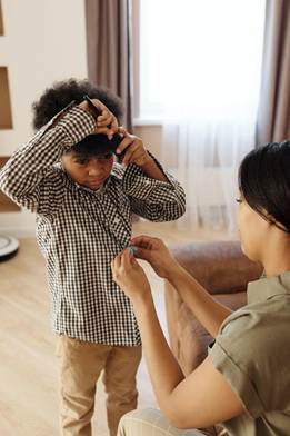A child with curly hair wearing a checkered shirt is getting assistance from an adult in getting dressed. The setting is in a bright, cozy room.