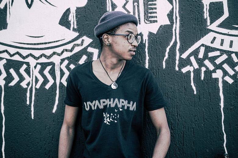 A young man wearing glasses and a beanie, standing against a wall adorned with abstract designs, dressed in a black t-shirt with the word 'UYAPHAPHANI' printed on it.
