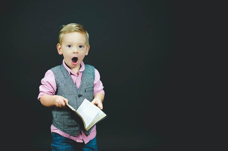 A surprised young boy holding an open book, wearing a vest and a pink shirt against a dark background.