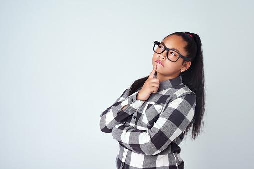 A thoughtful young girl in a plaid shirt with glasses, contemplating deeply with her finger on her chin and a slight smile.