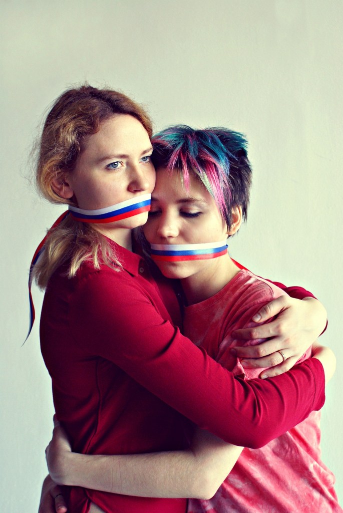 Two women embrace, with colored ribbons around their mouths, conveying a sense of connection and emotion.